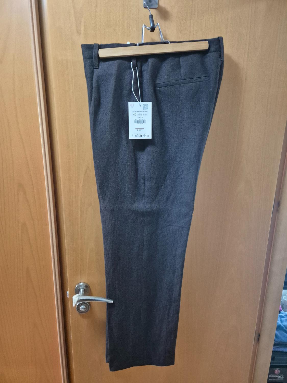 Grey trousers hanging on a wooden hanger over a light wooden door. Visible label with product details.