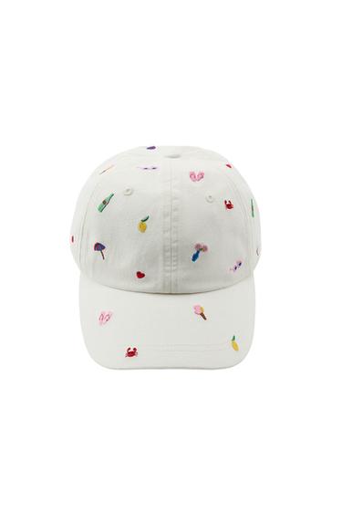 Front view of an ecru-coloured cap with small, multi-coloured embroidered designs of a lemon, ice cream, parasol, crab and other decorative motifs.
