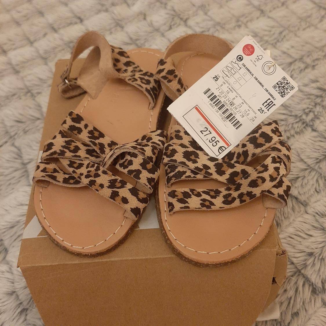 Παιδικά πέδιλα με animal print λεοπάρ πάνω σε καφέ κουτί. Ορατή ετικέτα με τιμή 27,95 €. Γκρι φόντο χαλιού.