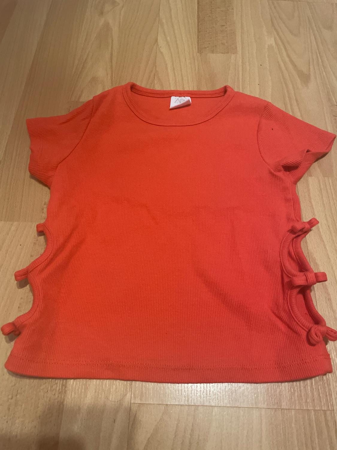 Red short-sleeved T-shirt with ties at the sides, on a light wooden floor. Visible label on the collar.