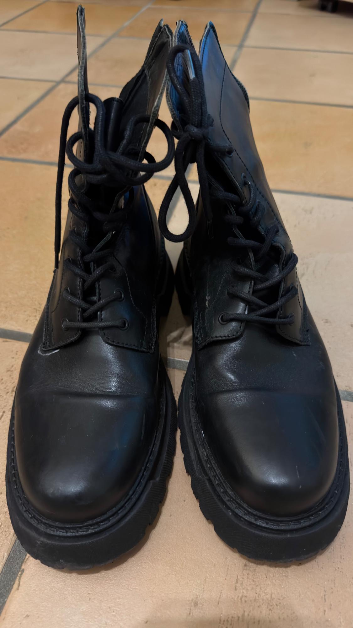 Black leather lace-up boots on a brown tiled floor. The image shows a front-on view of the boots.