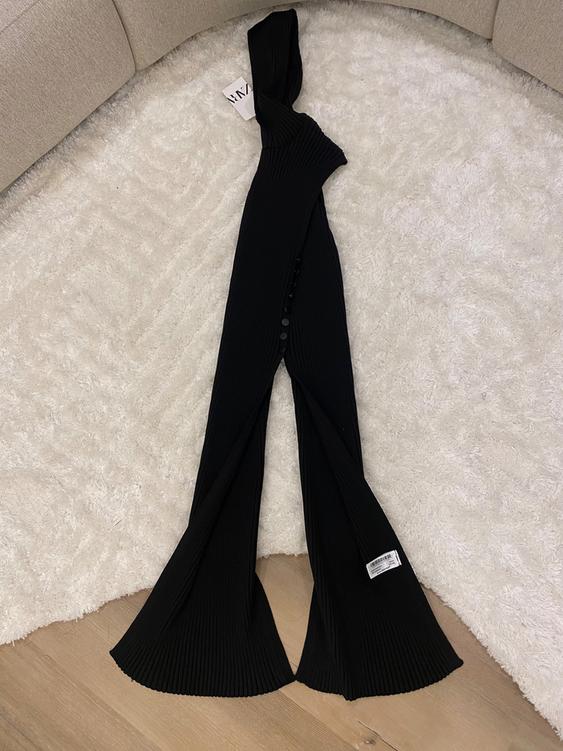 Black knit flared pants on a white rug, with a visible tag on top. Beige sofa background.