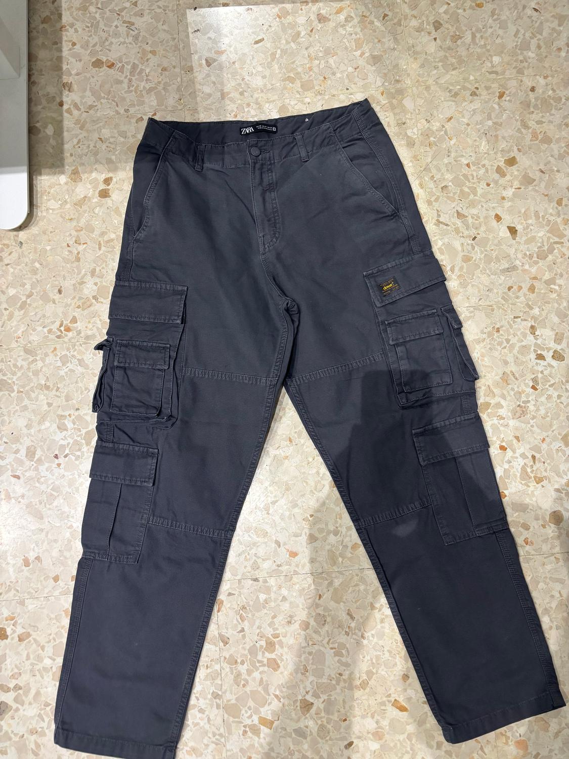 Black cargo trousers with multiple pockets, a visible label on the right pocket, on a light tiled floor.