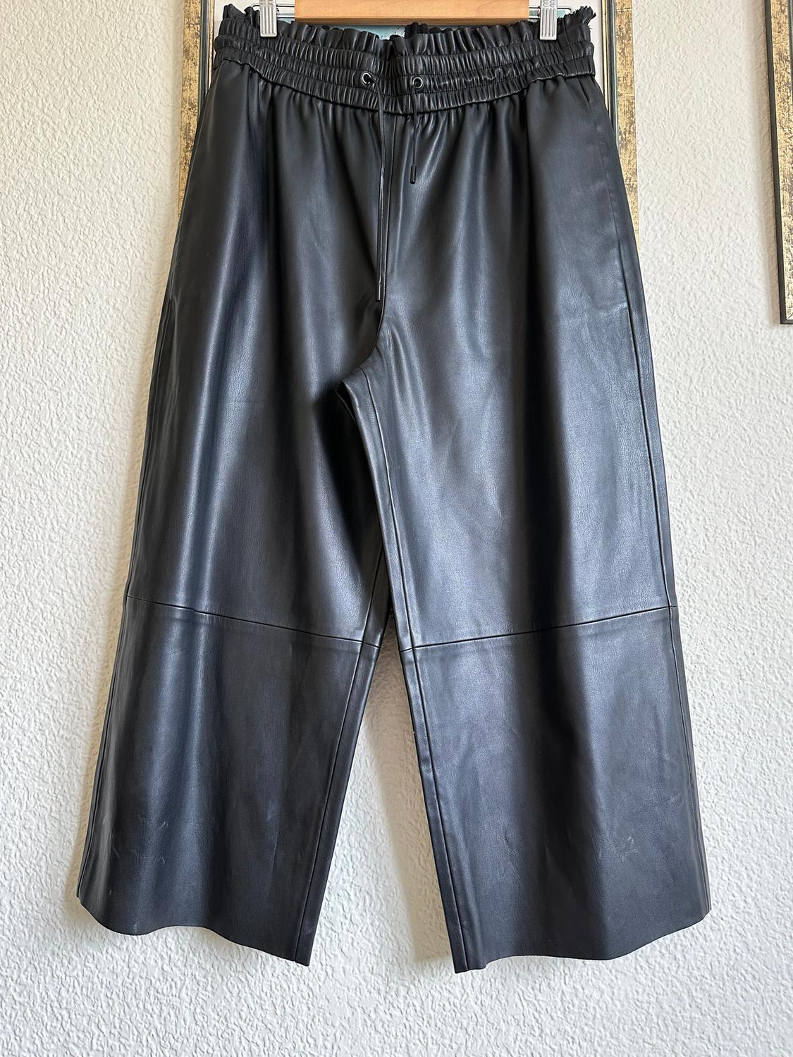 Black faux leather trousers hanging on a hanger, with an elasticated waist and adjustable drawstring, against a white wall background.