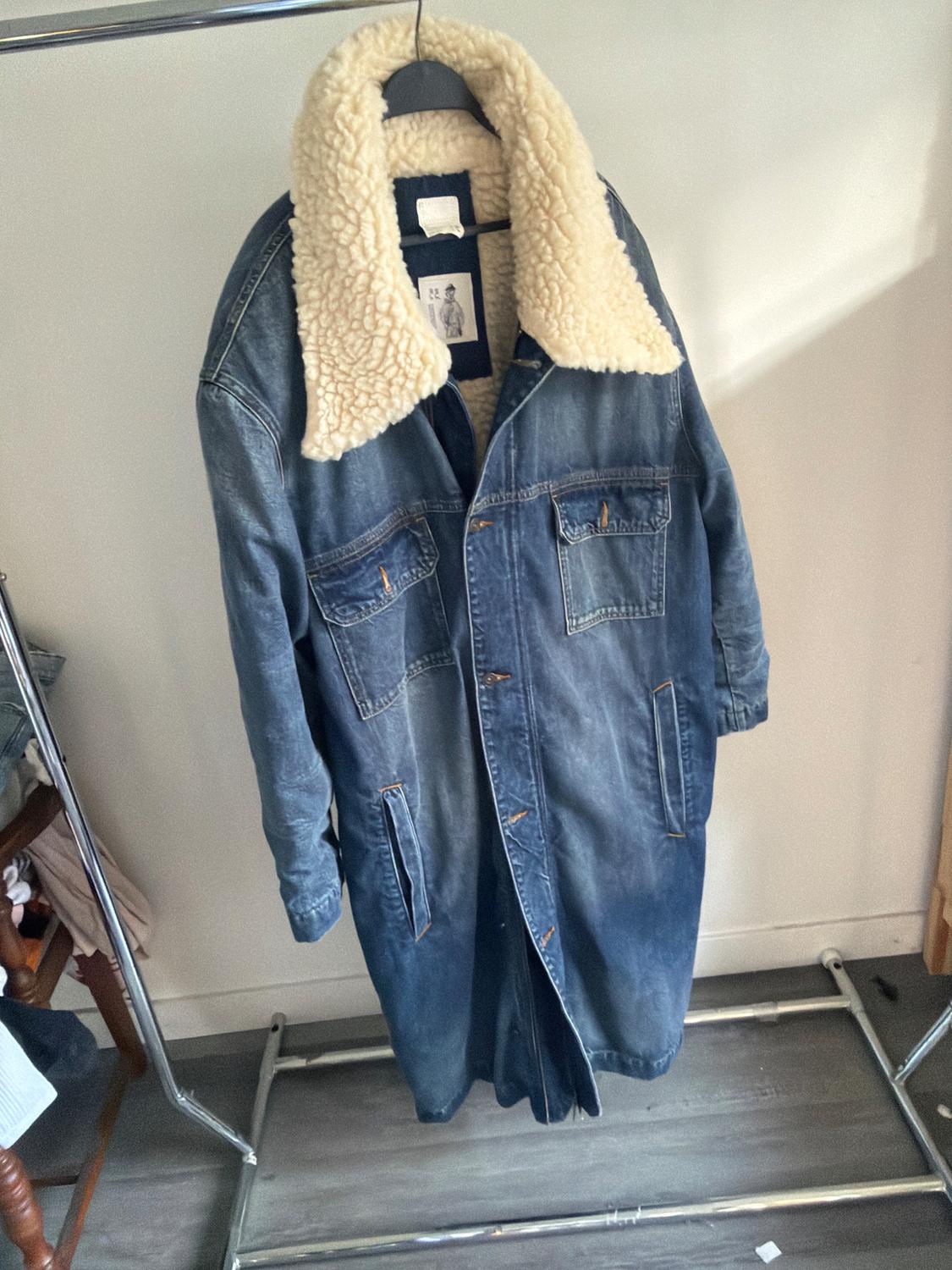 Blue denim coat with a white shearling collar, hanging on a metal clothes rack in a room with a wooden floor.