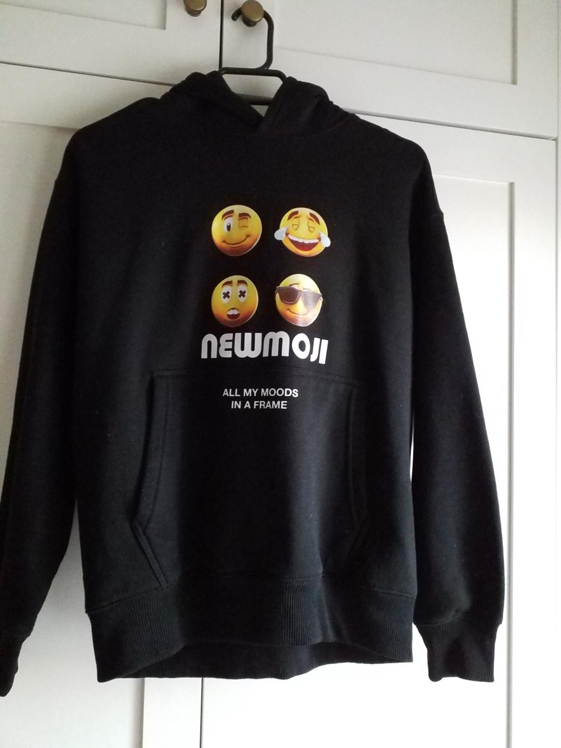 Black hoodie with a dropped hood, with emojis and text "NEWMOJI" and "ALL MY MOODS IN A FRAME" on the front, against a white wardrobe background.