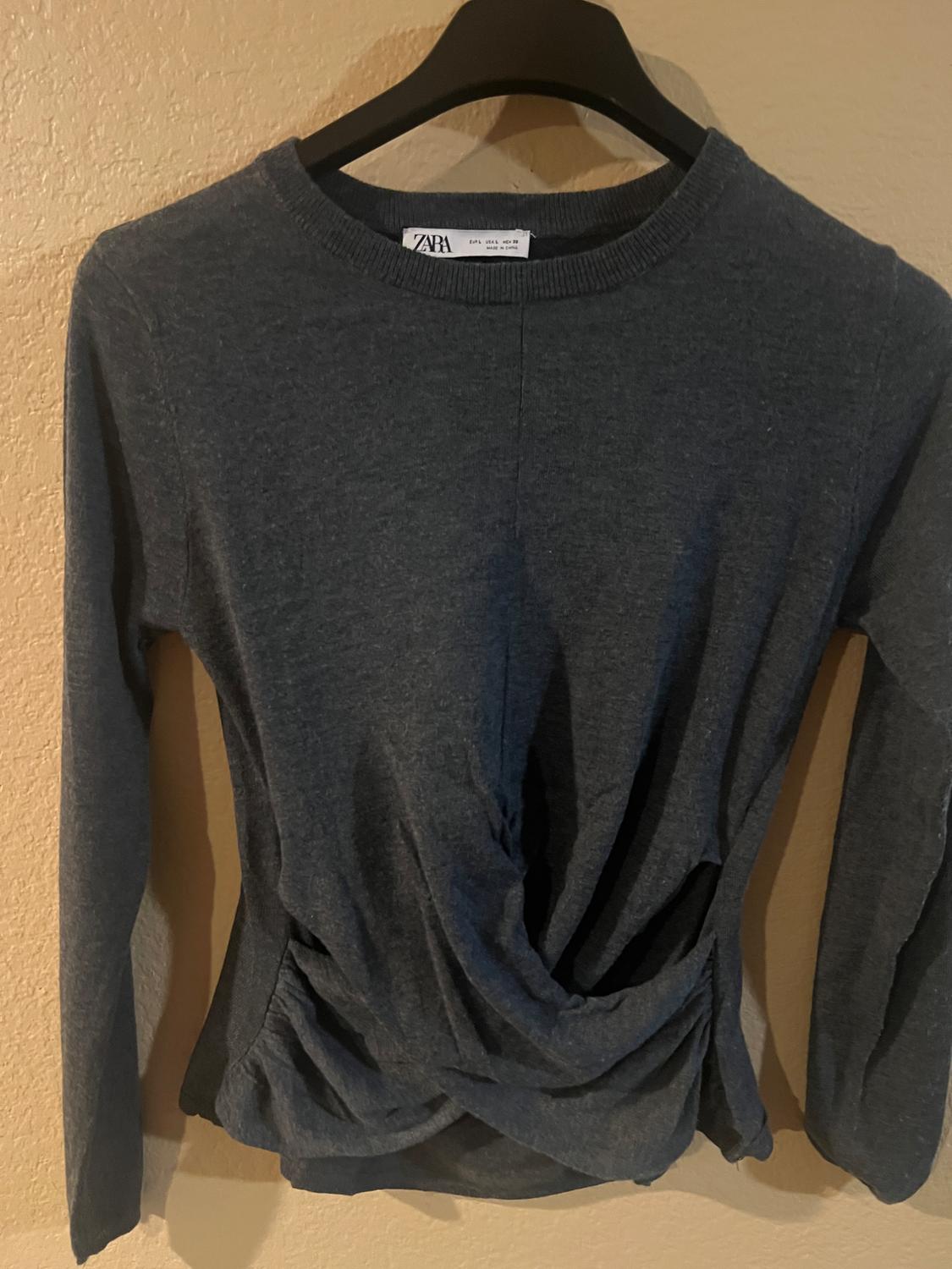Dark grey long-sleeved jumper with draped detailing at the waist, hanging on a black hanger against a beige wall.