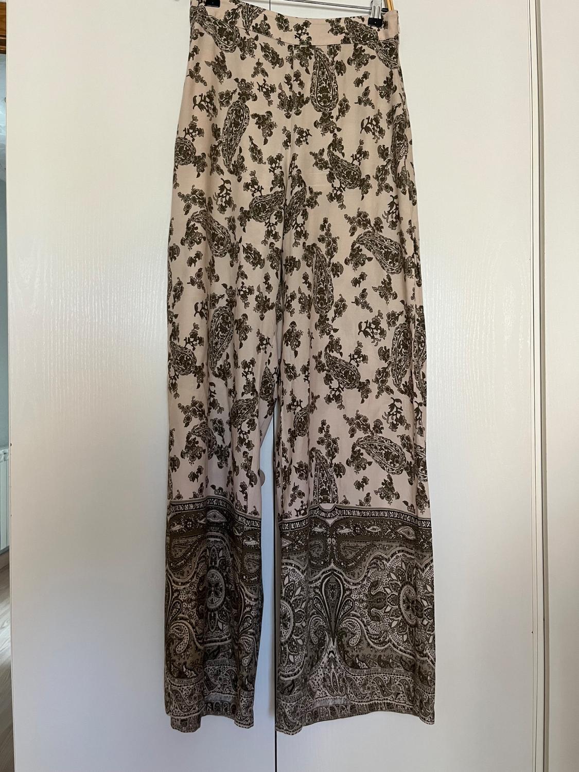 Beige wide-leg trousers with a dark brown paisley and floral print, with a border at the hem, hanging on a hanger on a white door.