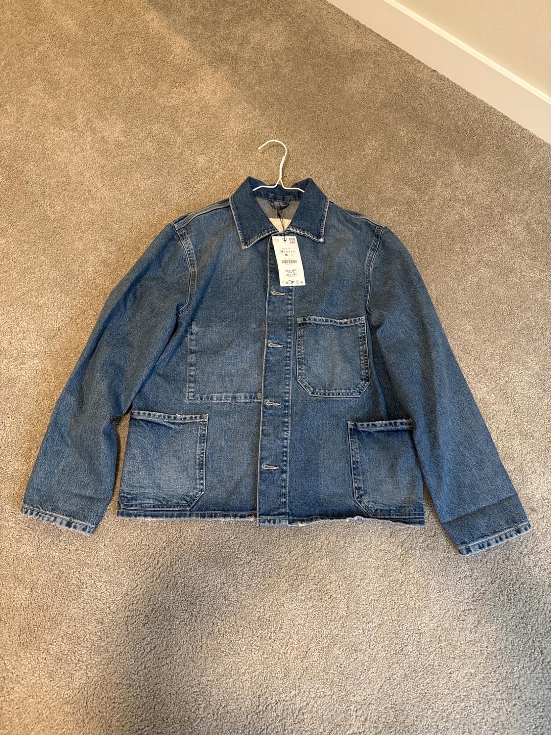 Blue denim jacket with large pockets, hanging on a hanger over a beige rug. Tag visible on the collar.