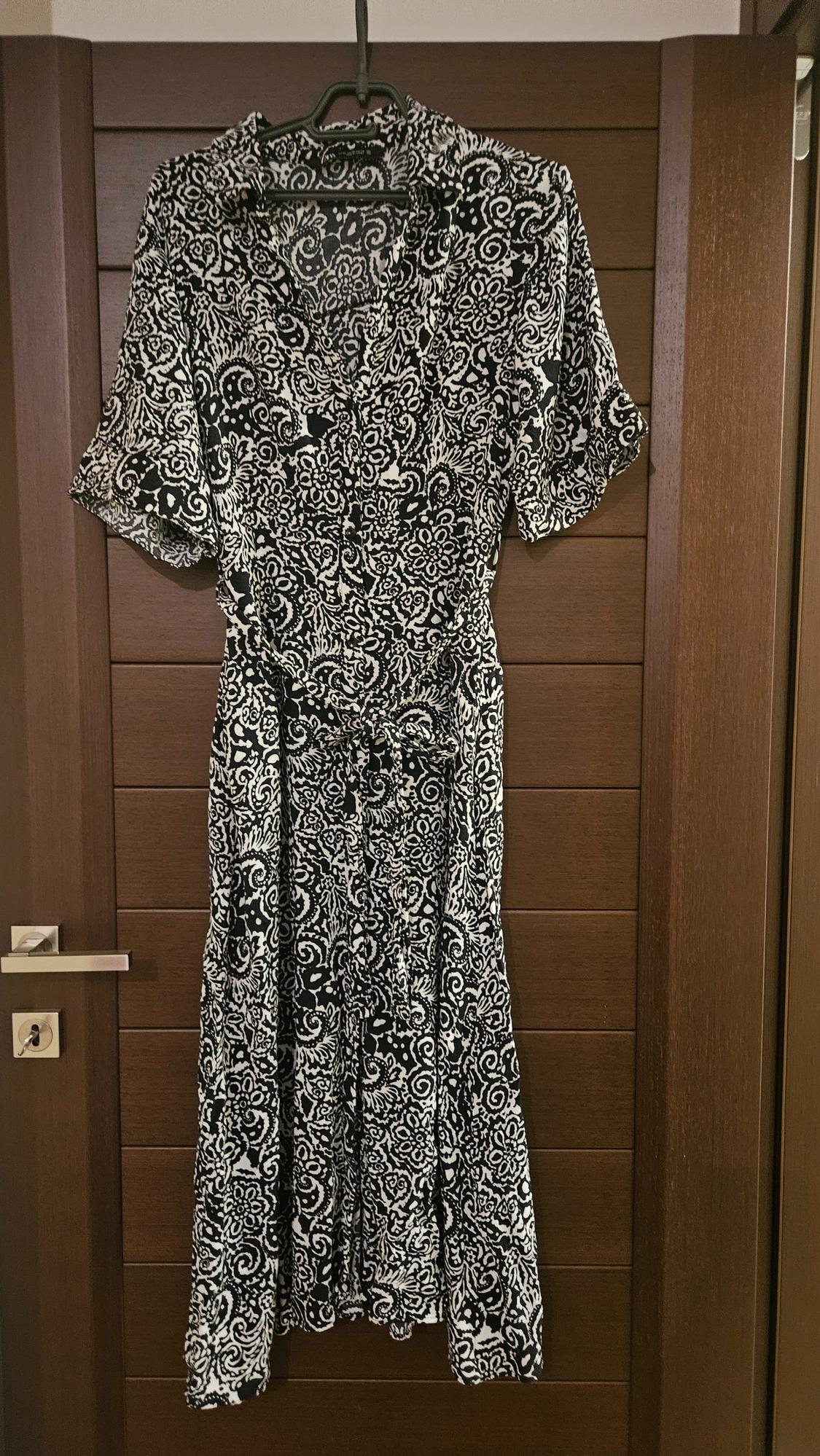 Dress hanging on a wooden door, with a black and white floral and geometric pattern, short sleeves and a belt.