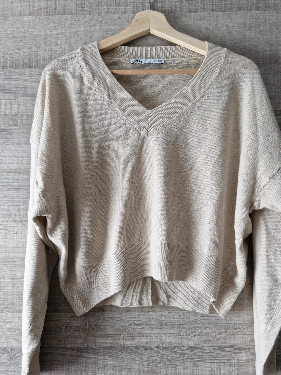 Beige knitted jumper with a V-neck hanging on a wooden hanger against a background of planks. Text: ZARA.