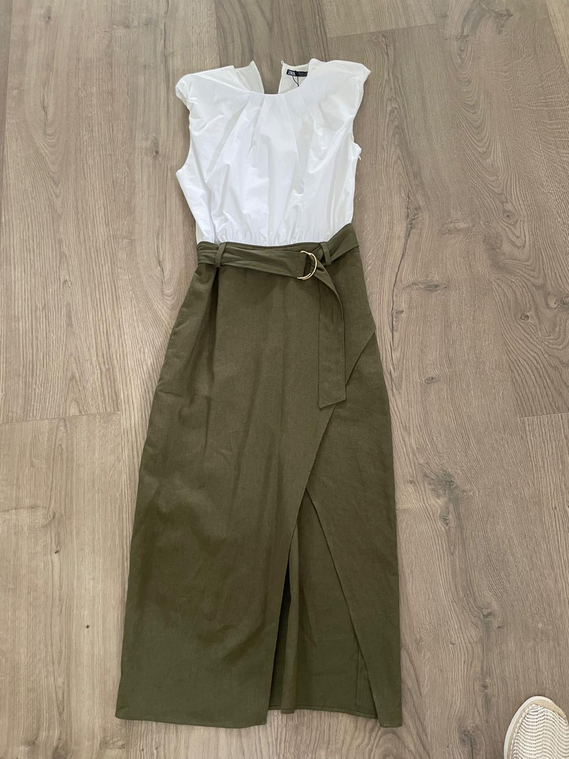 Dress on a wooden floor. White sleeveless top with a gathered neckline and an olive green wrap skirt with a slit and belt.
