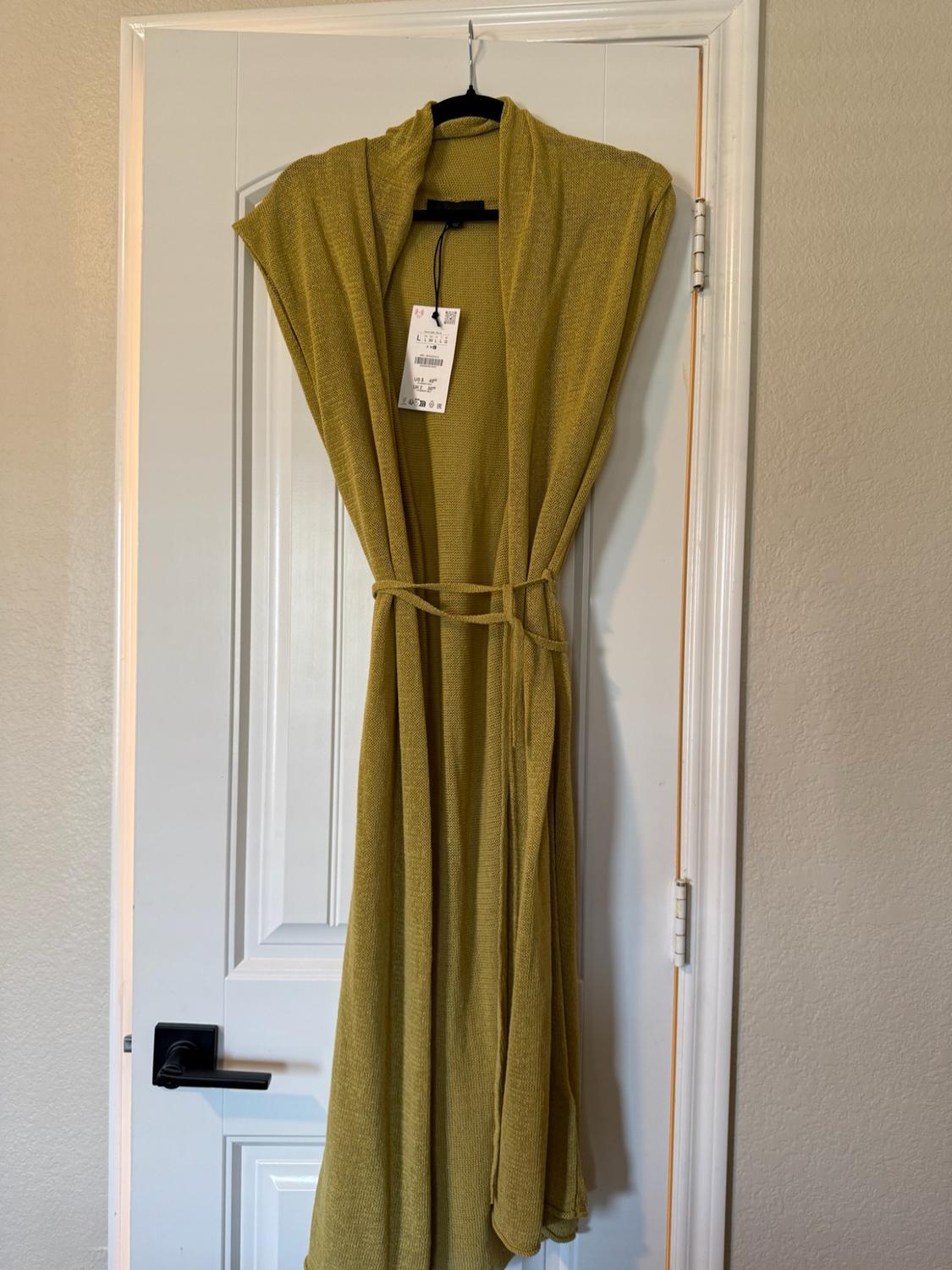 Mustard-coloured long knitted waistcoat with belt and tag, hanging on a hanger over a white interior door.