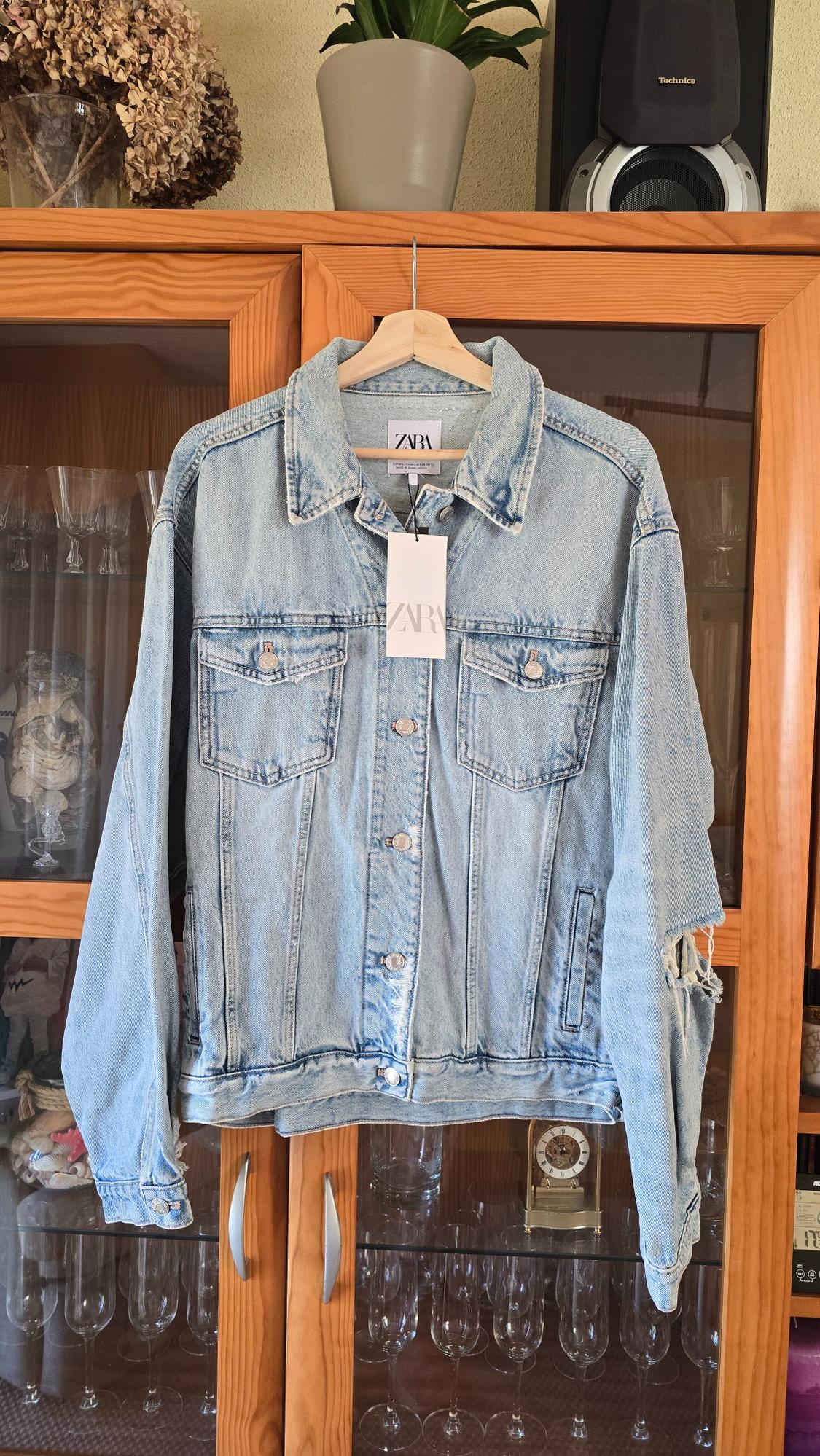 Light blue denim jacket hanging on a wooden hanger, with a "ZARA" label, on a wooden piece of furniture with crystal glasses.