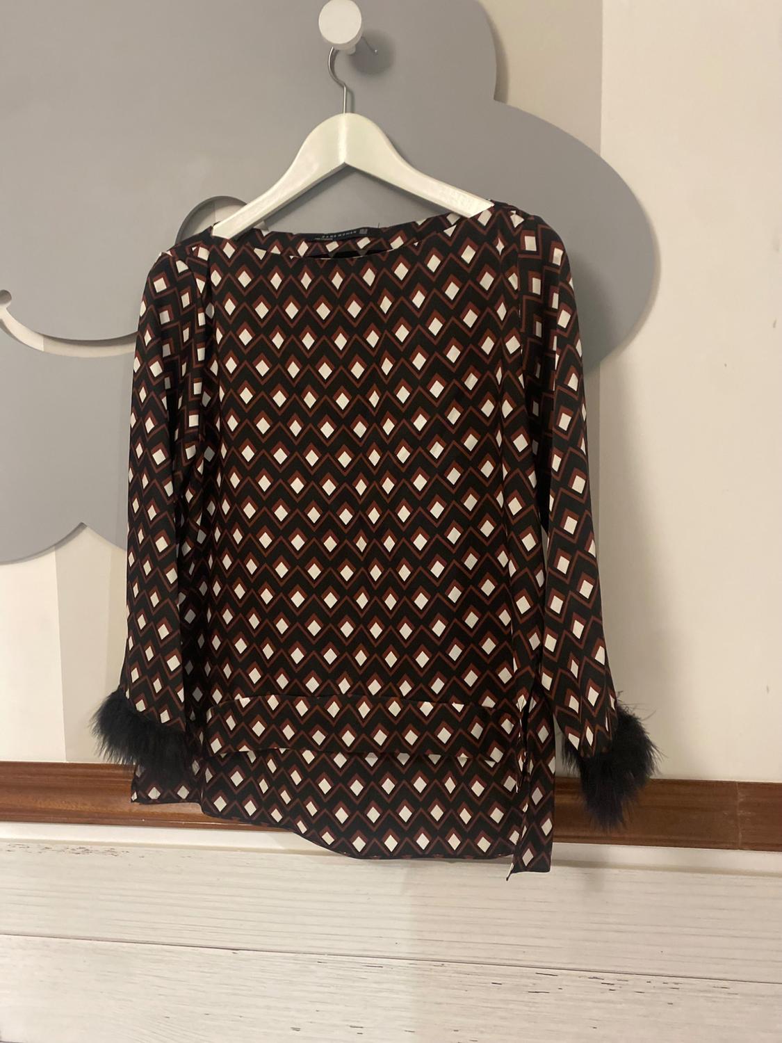 Long-sleeved blouse with a geometric print in brown and white tones, black feather details on the cuffs, hanging on a white hanger.