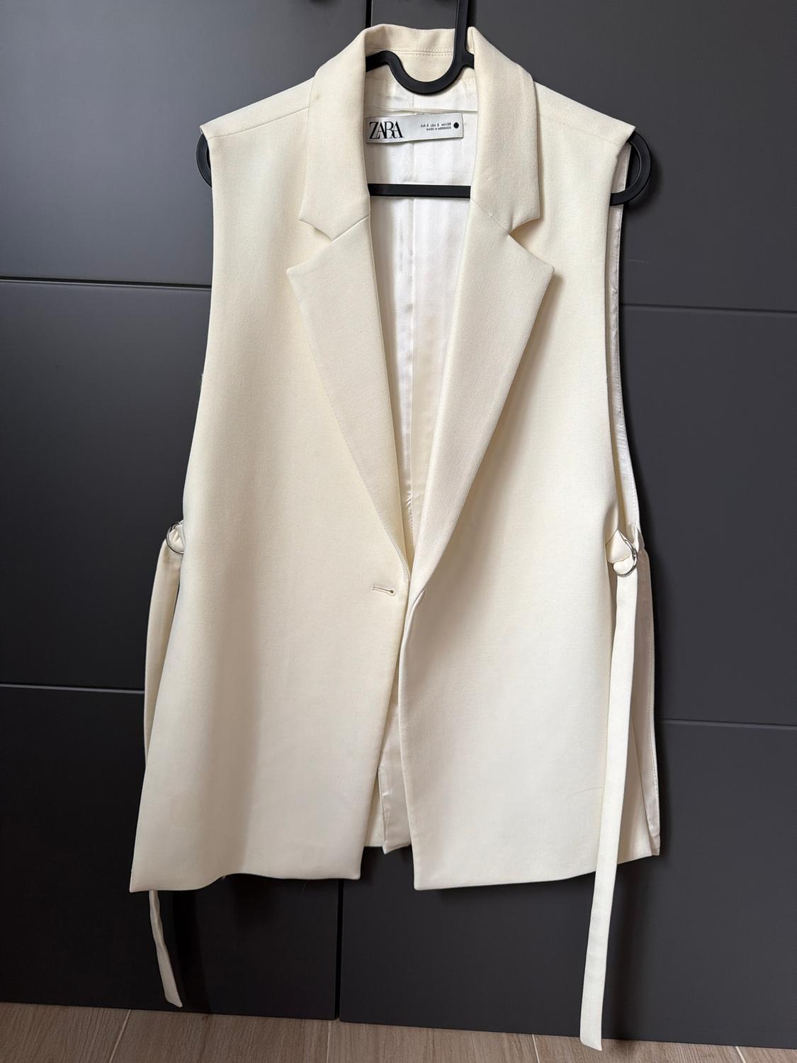 Cream sleeveless waistcoat hanging on a black hanger, with a visible Zara tag, against a dark background.