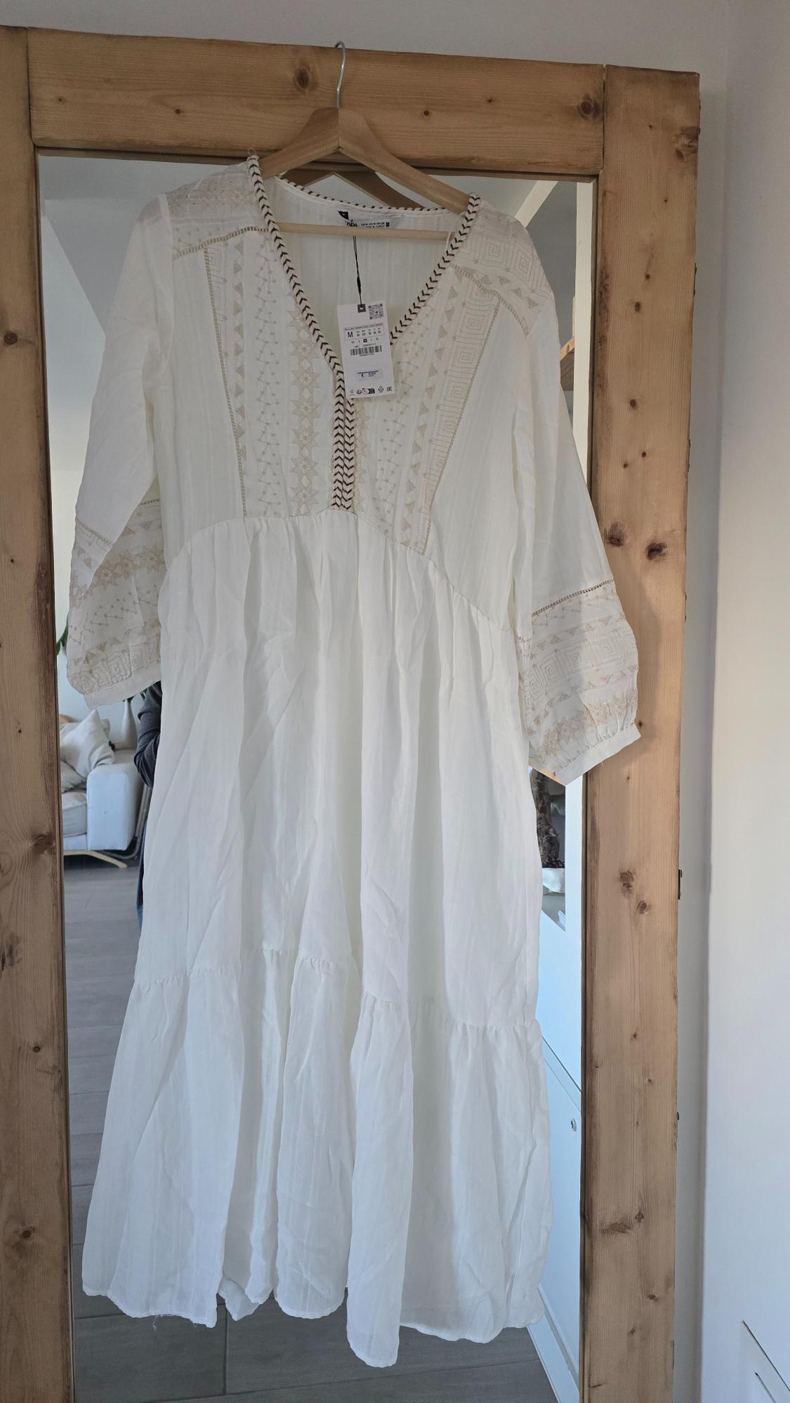 White embroidered dress hanging on a wooden hanger. Reflection of a sofa in the mirror behind. Label visible on the neckline.