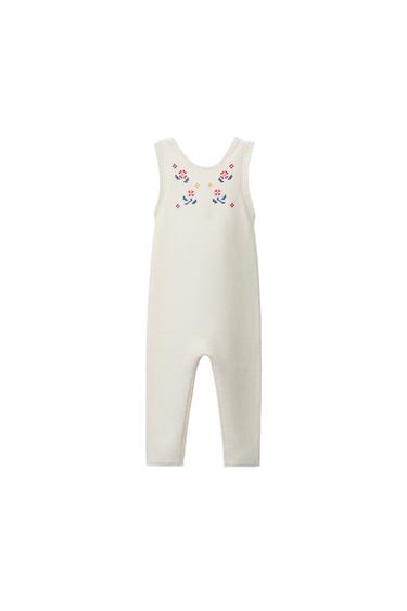 Front view of a baby's bib in an unbleached colour with multicoloured floral embroidery on the chest and wide straps.