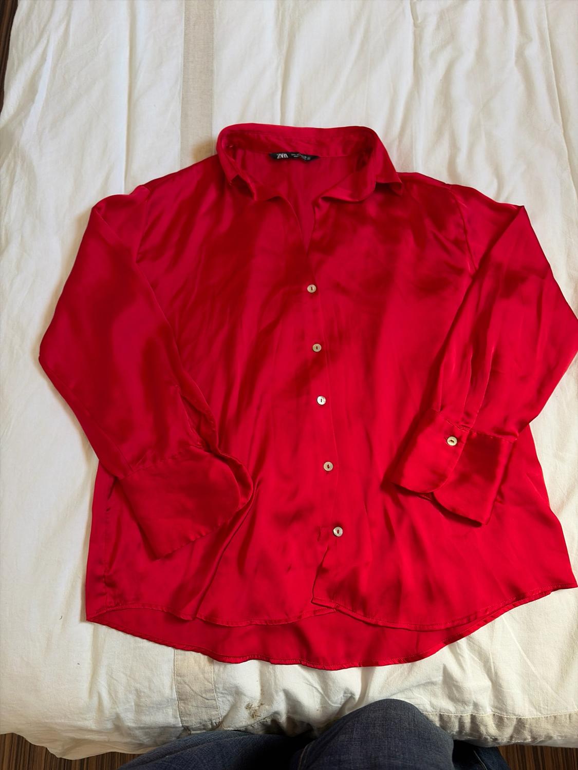 Red long-sleeved button-up shirt in a shiny fabric, laid out on a white sheet. A pair of jeans is visible at the bottom edge.