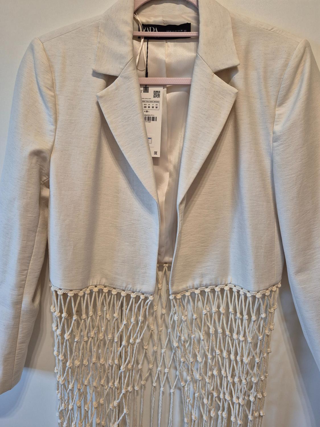 White jacket with lapel collar and fringed mesh hem, hanging on a hanger. Text on label: ZARA, 28 M M M, EAC.