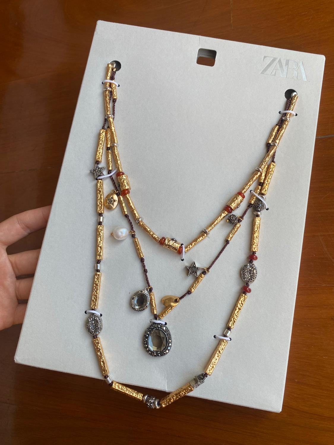 Hand holding a white piece of cardboard with the text ZARA on wood, showing a multi-layered necklace with gold beads and pendants.