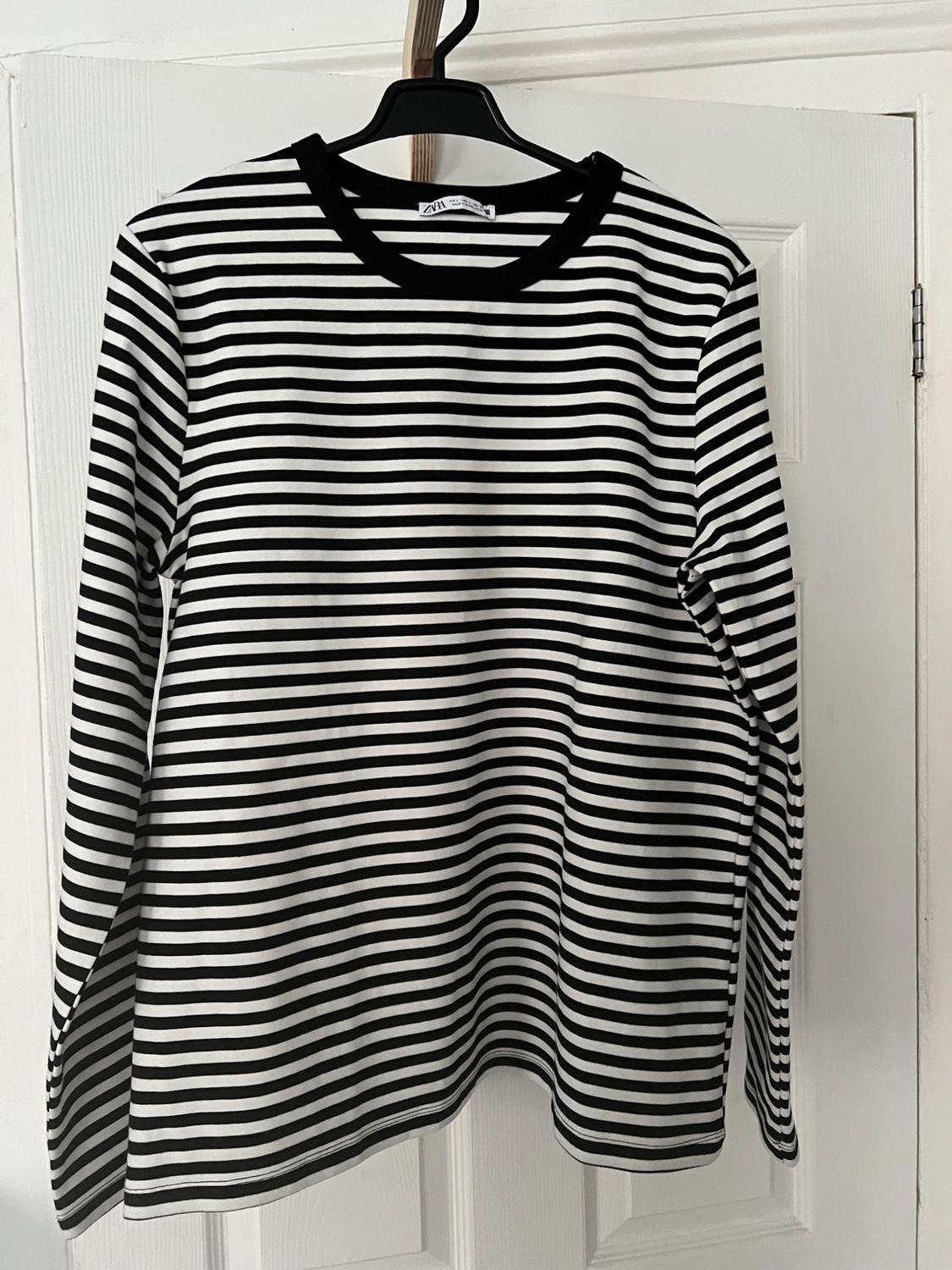 Black and white horizontally striped long-sleeved t-shirt with a black collar, hanging on a white door. Label with the text Zara.