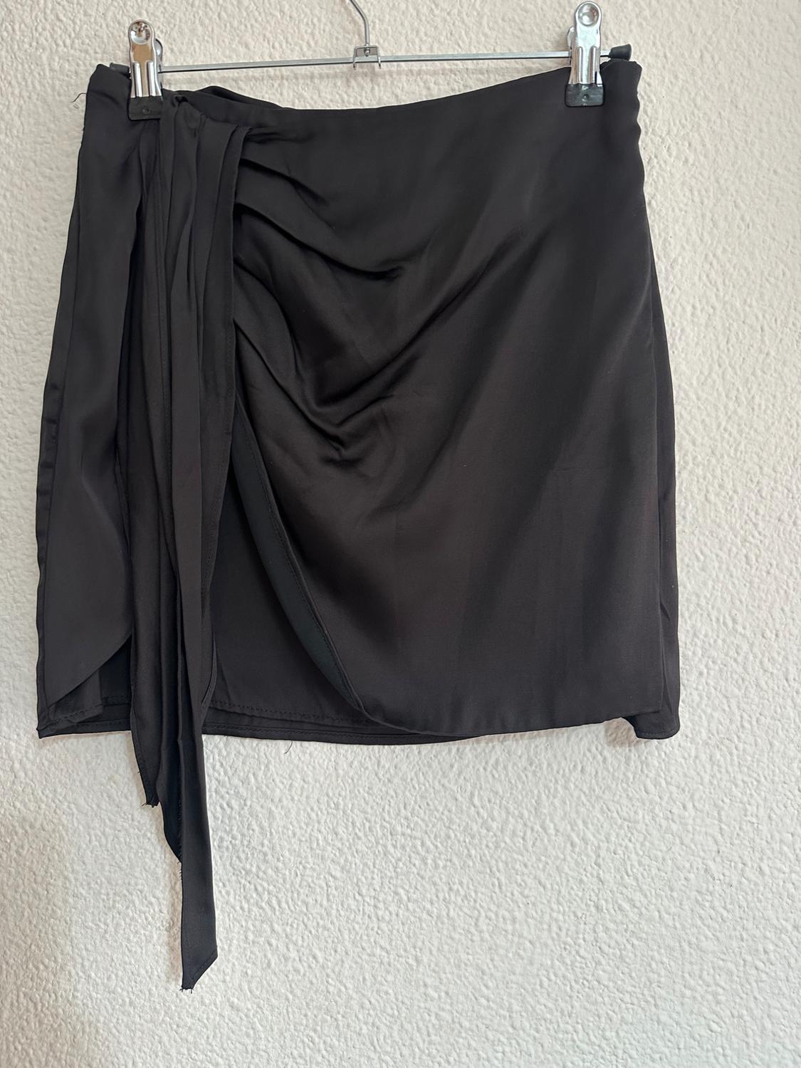 Short black skirt with draping and a side tie, hanging on a hanger against a white wall.