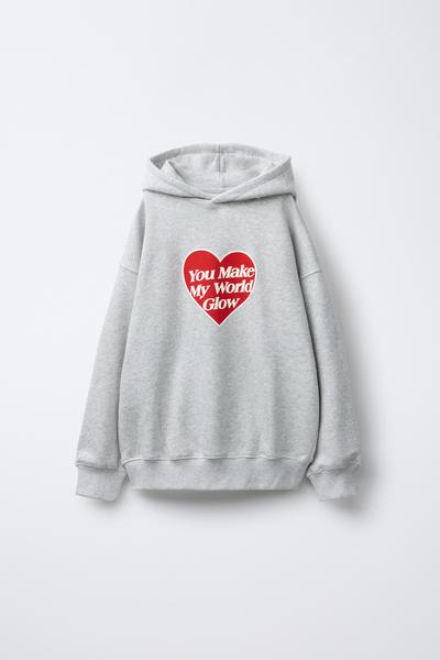 Vorderansicht eines grauen Kapuzenpullovers mit dem Text „You Make My World Glow“ in einem roten Herz auf der Brust. Rippbündchen.