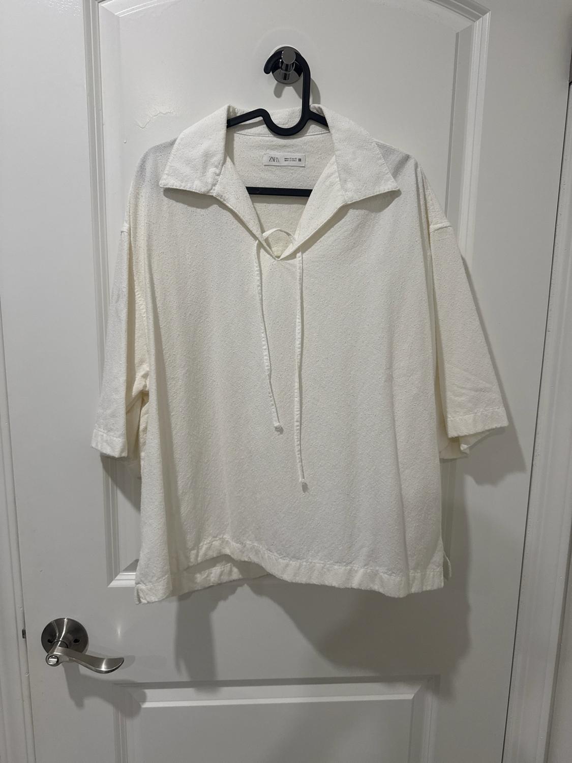 Textured white short-sleeve shirt with a collar and drawstrings, hanging on a black hanger over a white door. ZARA label.