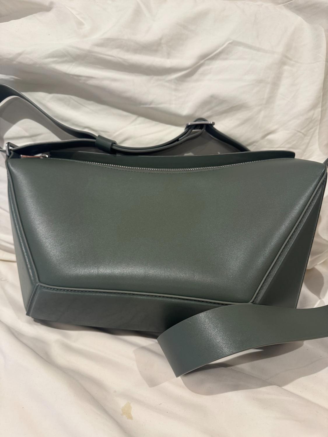 Dark grey leather bag with a geometric design on a wrinkled white fabric background. Wide strap and visible top closure.