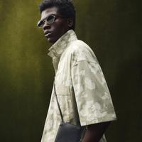 Image 2: Model wearing sunglasses and a beige floral shirt, carrying a crossbody bag. Dark green background, modern and relaxed style.