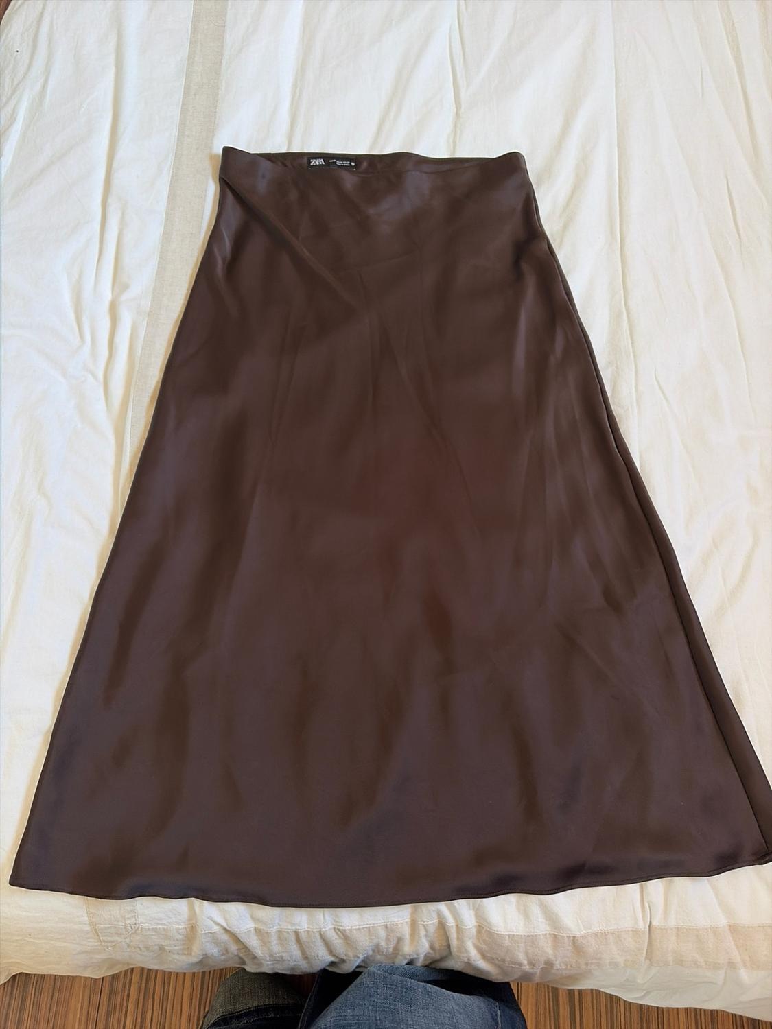 Dark brown satin midi skirt laid out on a white sheet. ZARA label at the waist. Relaxed atmosphere.
