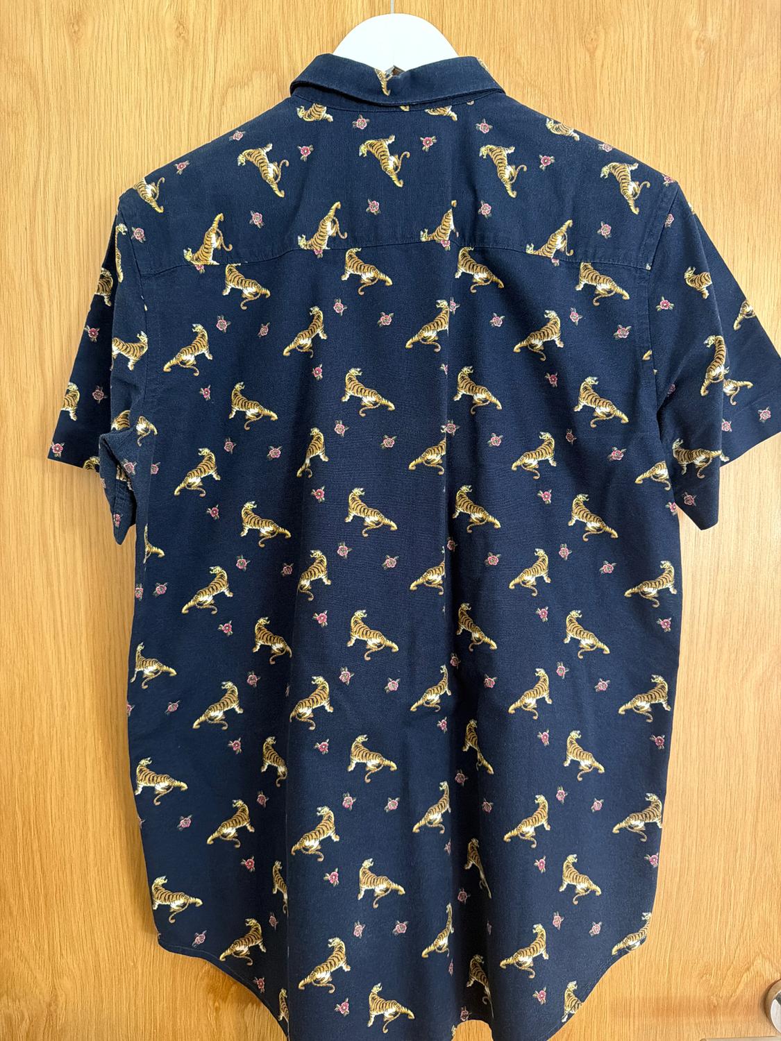 Navy blue shirt with yellow tiger print and small floral motifs, hanging on a hanger against a light wood background.