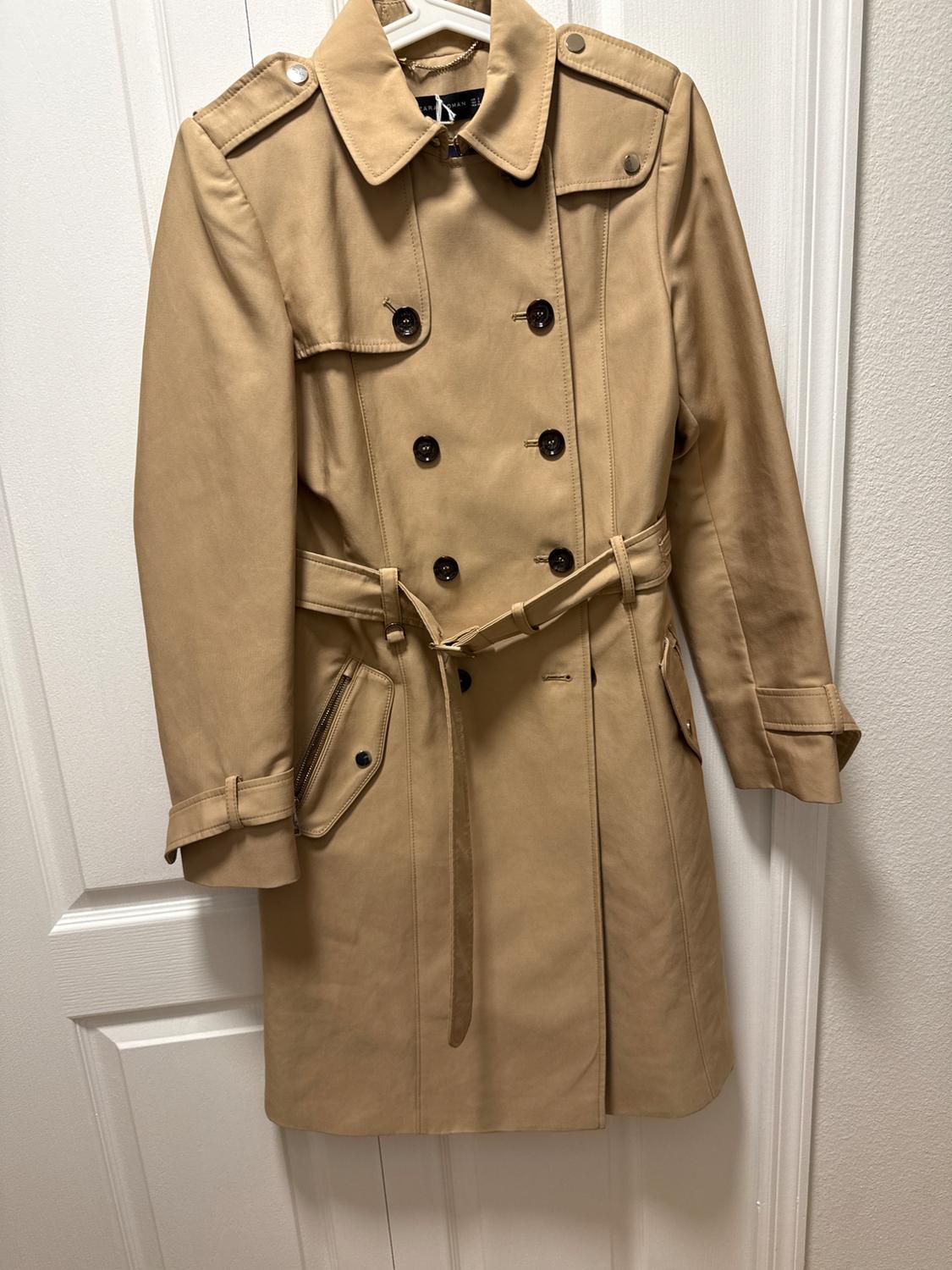 Beige trench coat hanging on a white door, with double-breasted closure and matching belt. Classic and elegant style.