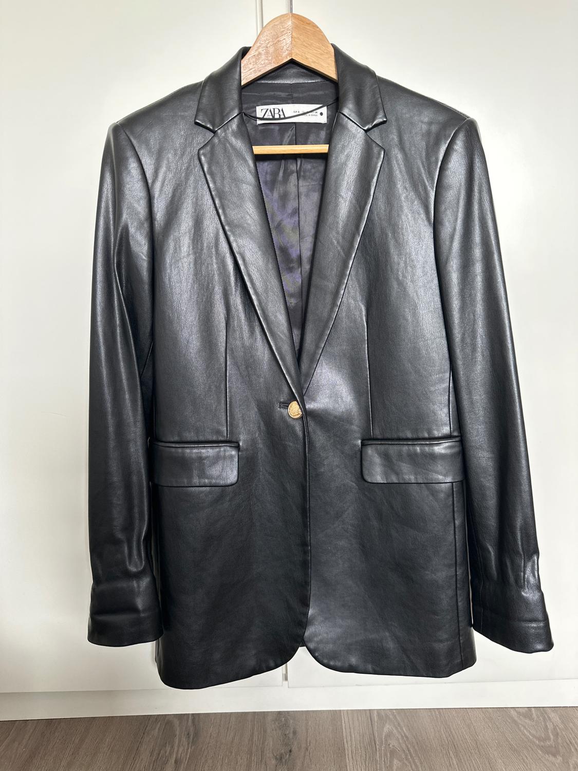 Black jacket hanging on a wooden hanger, with classic lapels and a gold button. White background and wooden floor.