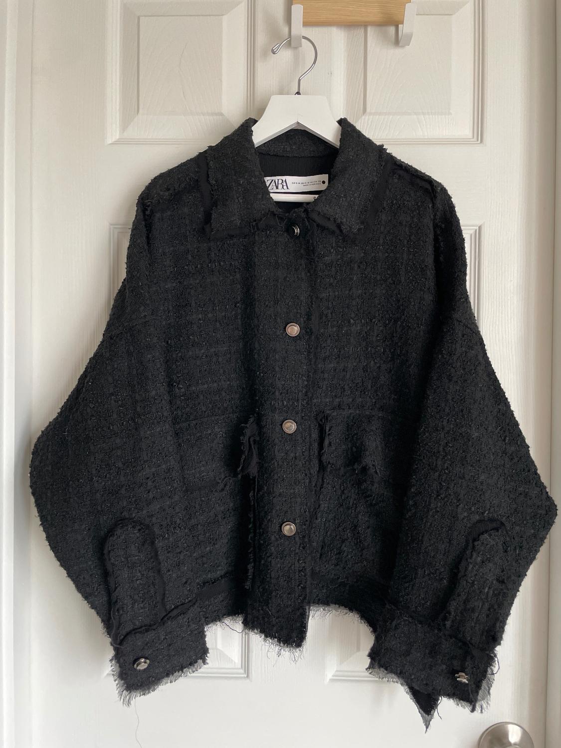 Black textured fabric jacket hanging on a white hanger over a white door. Visible label with the text "ZARA".