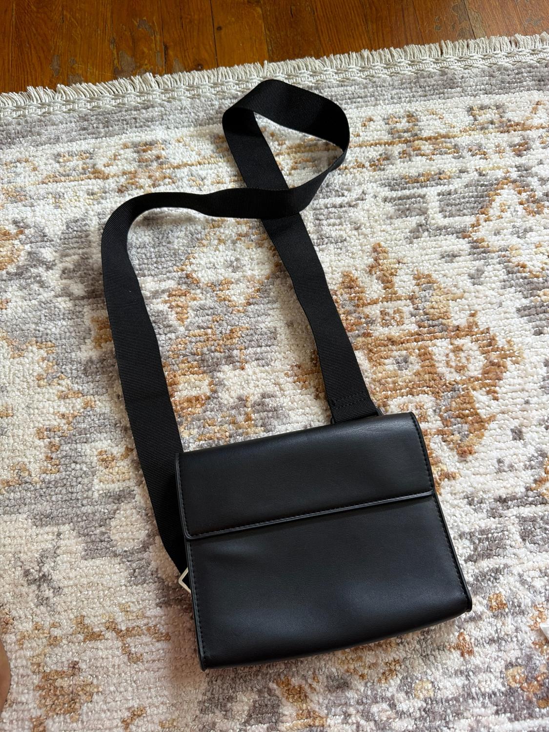 Black flap bag with a wide strap, resting on an earth-toned patterned rug and a wooden floor.