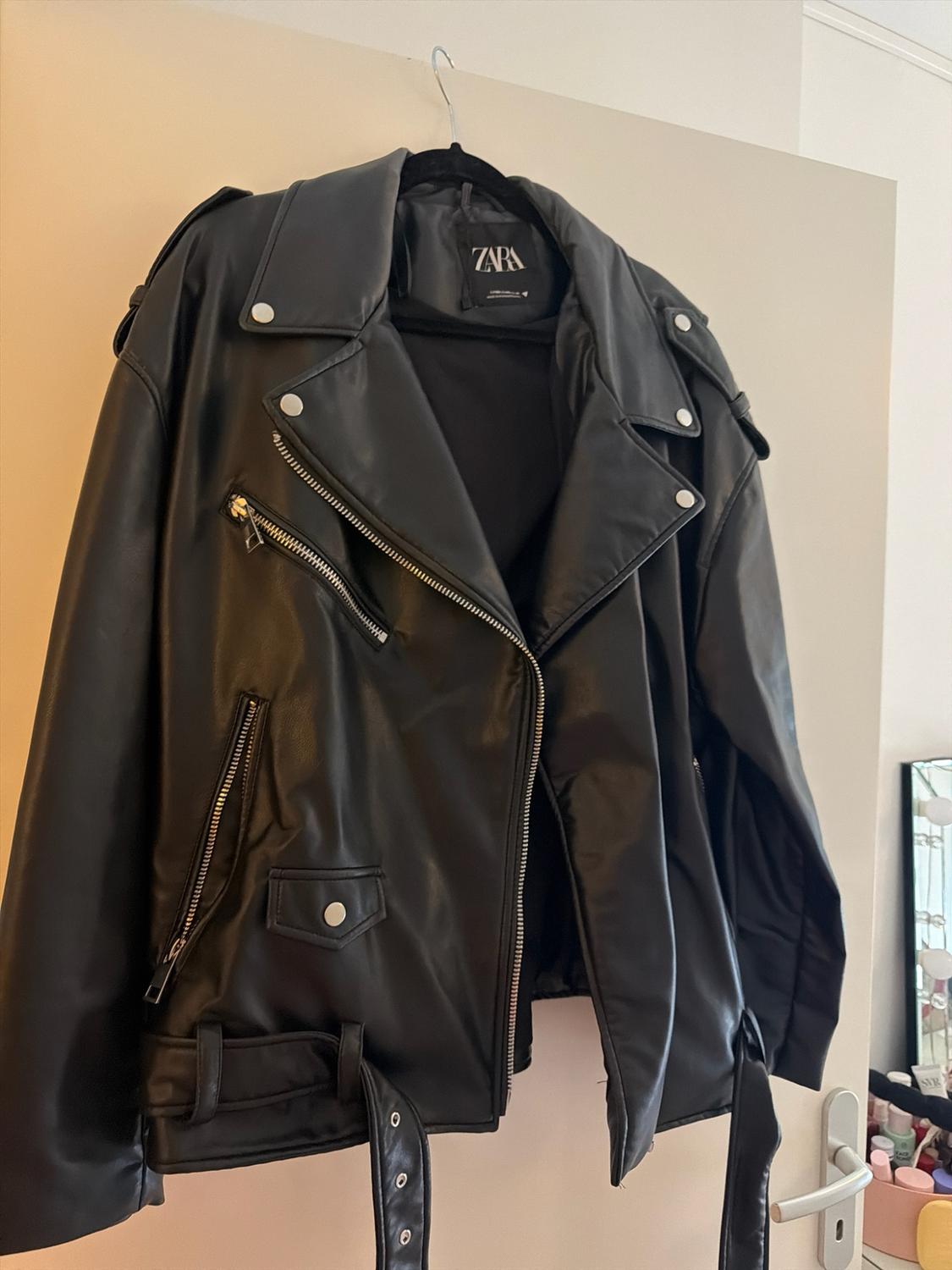 Black biker-style jacket with zips and a belt, hanging on a coat hanger on a white door. Inner label with the text ZARA.