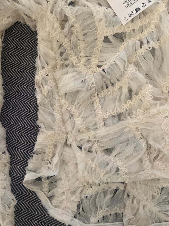 White fabric with feather texture and zigzag details on a black and white herringbone pattern background. Label visible in the corner.