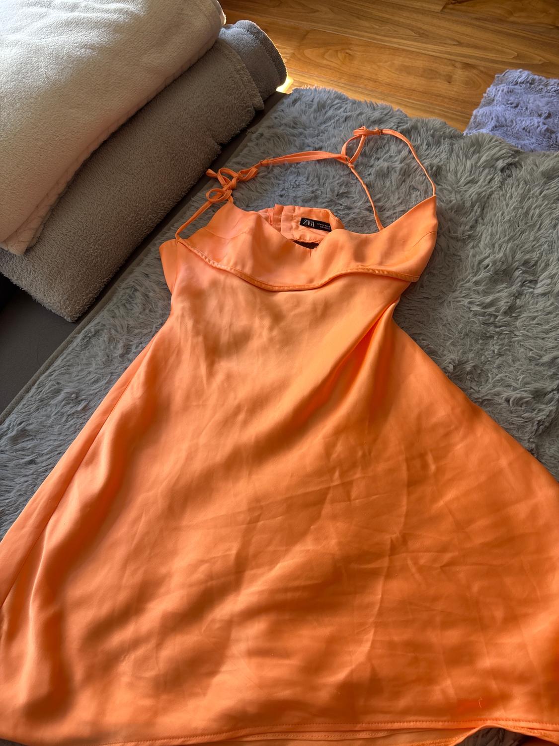 Πορτοκαλί σατέν φόρεμα με ετικέτα ZARA, πάνω σε ένα χνουδωτό γκρι χαλί δίπλα σε διπλωμένες πετσέτες σε ξύλινο πάτωμα.
