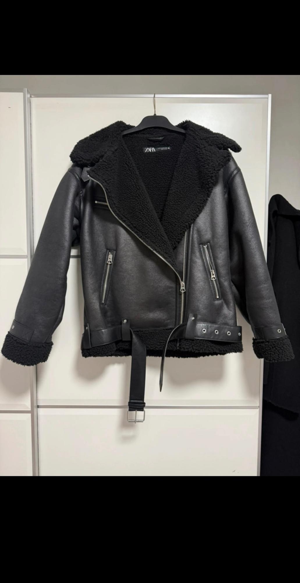 Black jacket with wool interior and loose belt, hanging on a hanger against a white background.