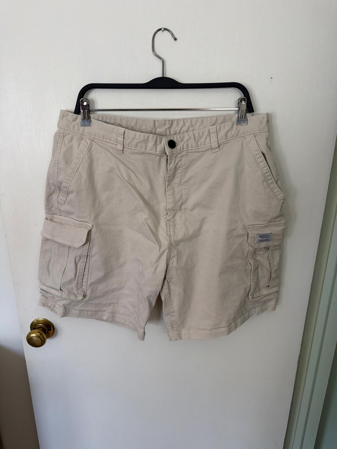 Beige cargo shorts hanging on a black hanger over a white door with a gold doorknob. They have side flap pockets.