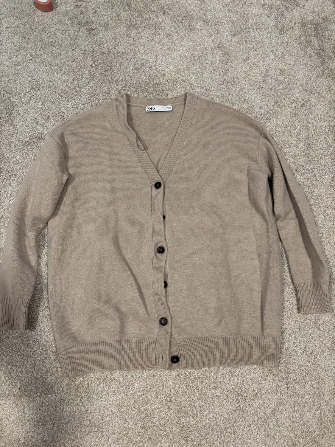 Beige long-sleeved cardigan with black buttons on a gray carpet. Visible label with text "ZARA".