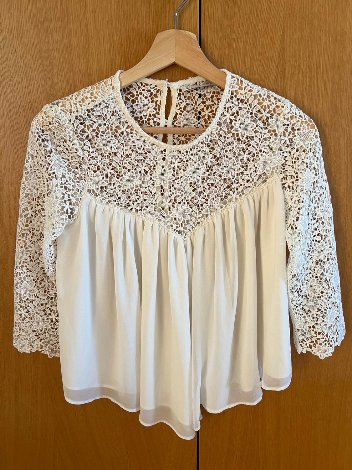 Romantic white blouse on a hanger against a wooden background. It features a yoke and floral lace sleeves, combined with a fluid, semi-sheer bodice.