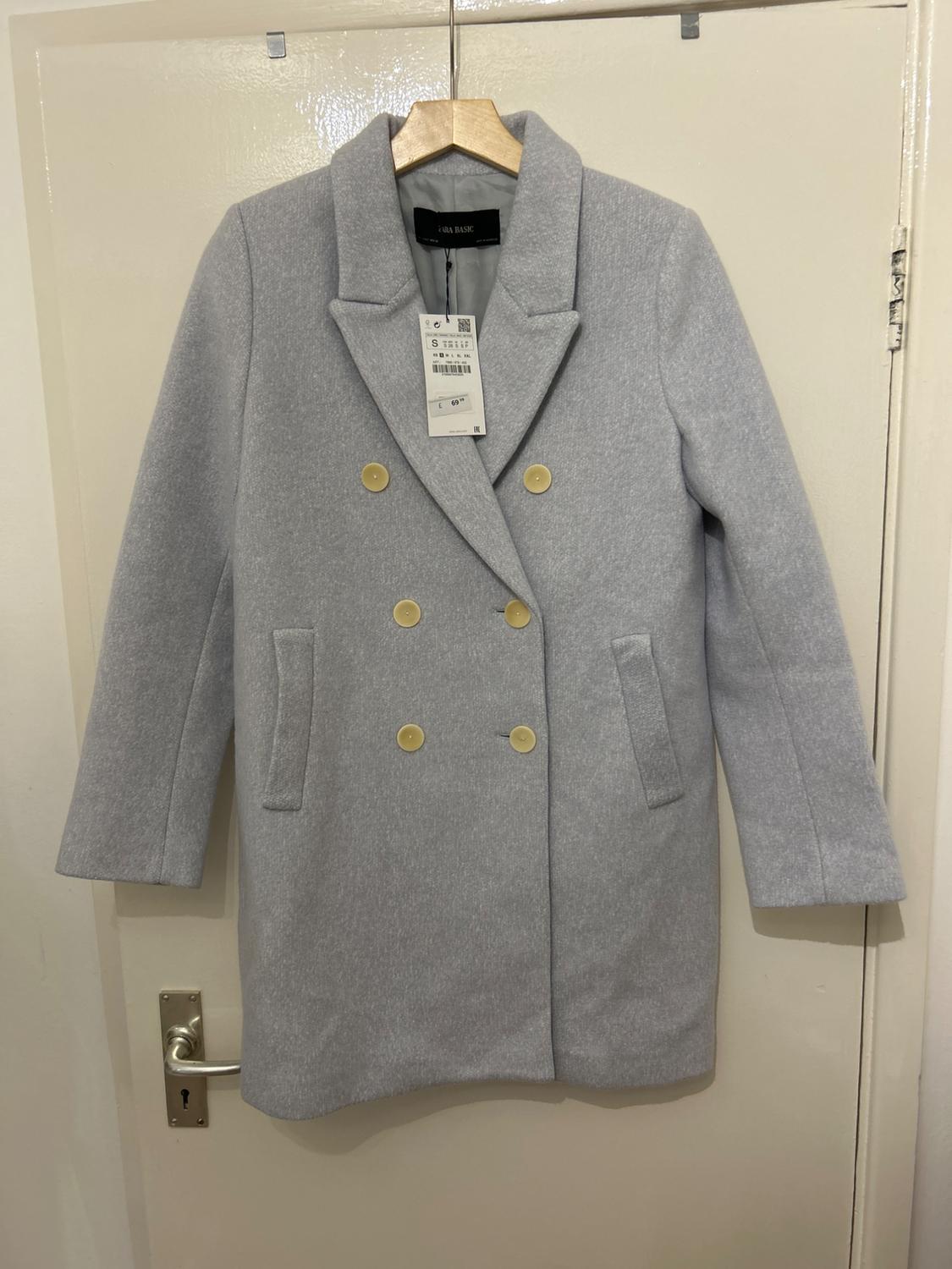 Light grey wool coat hanging on a door, with gold buttons and visible label.