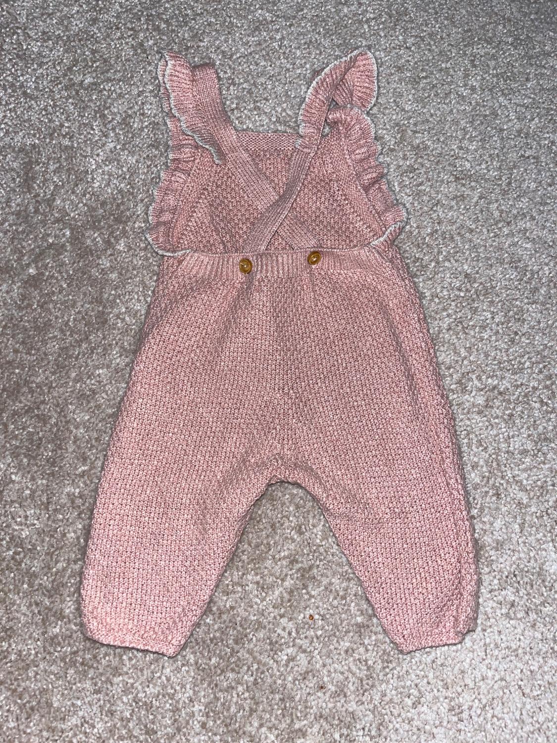 Knitted baby romper in pink with crossed straps and ruffles, on a grey rug.