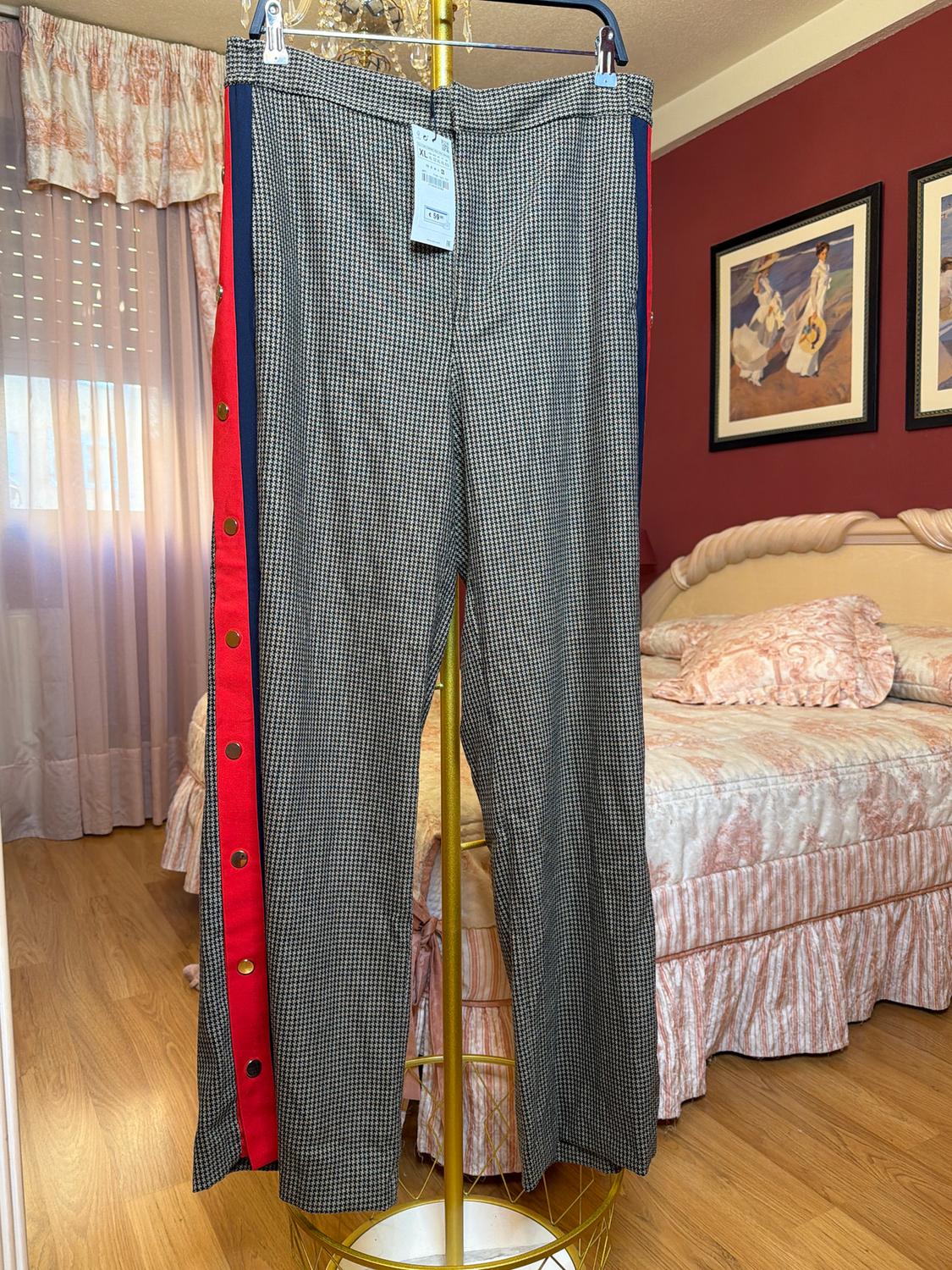Trousers with a checked pattern hanging on a gold hanger in a bedroom with pink curtains and a bed. Red detail with buttons on the side.