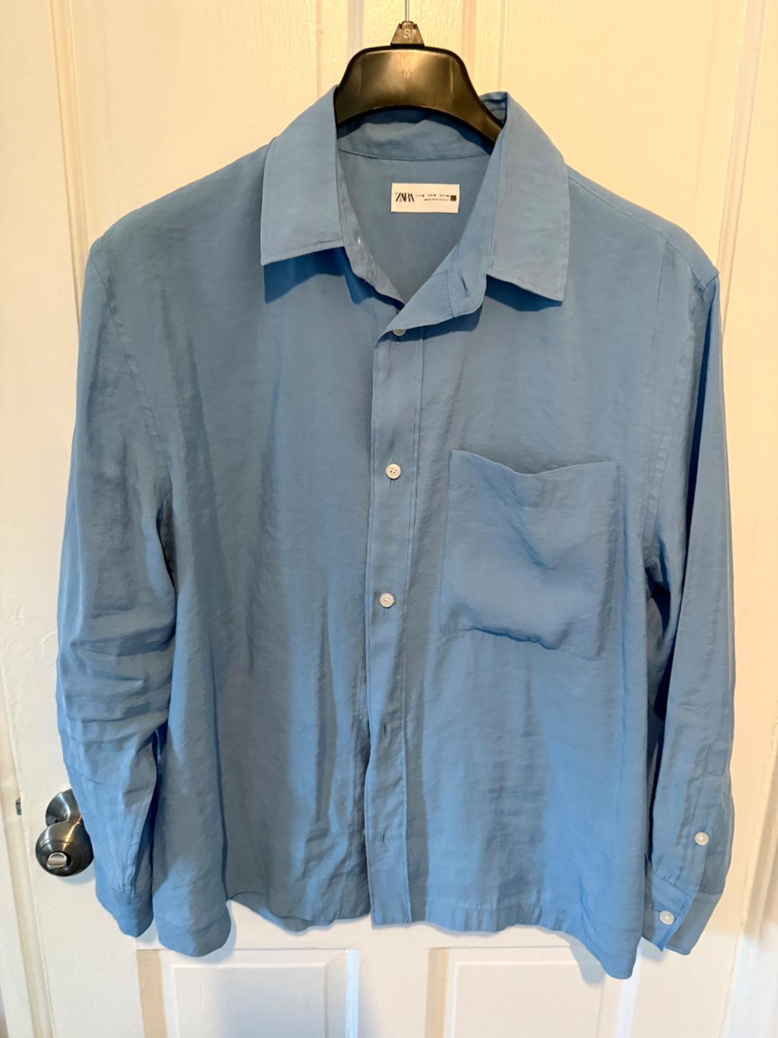 Light blue shirt hanging on a black coat hanger over a white door. It has a breast pocket and white buttons.