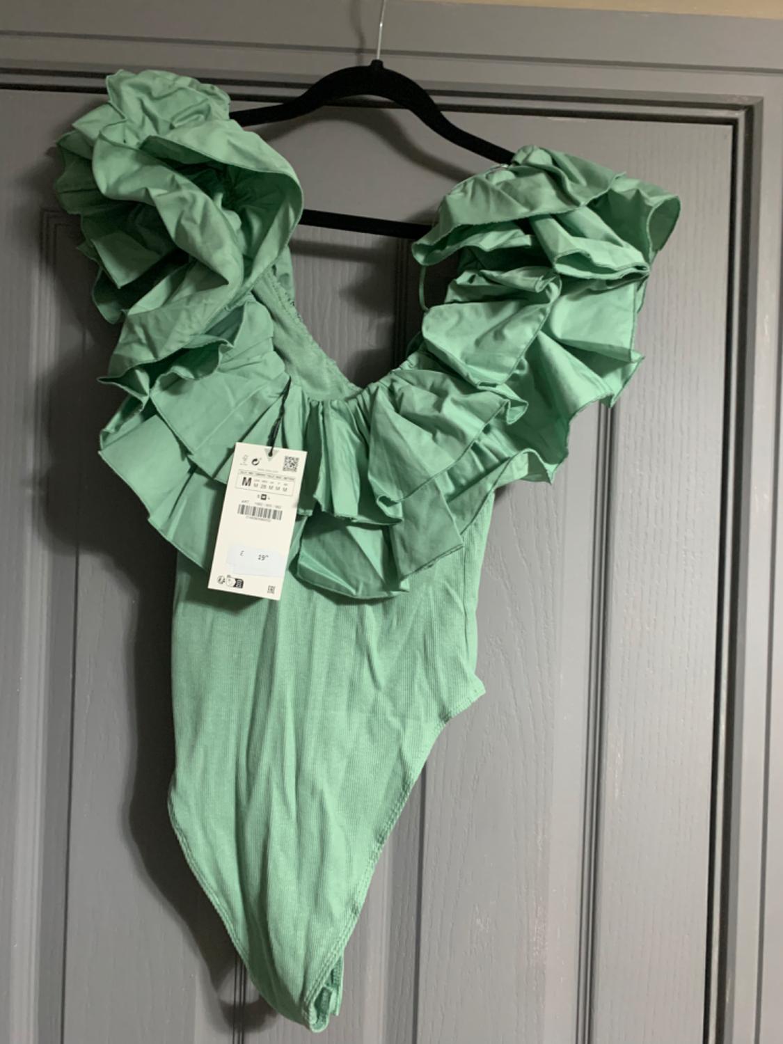 Green bodysuit with large ruffles on the shoulders, hanging on a black hanger over a grey door. Visible price tag.