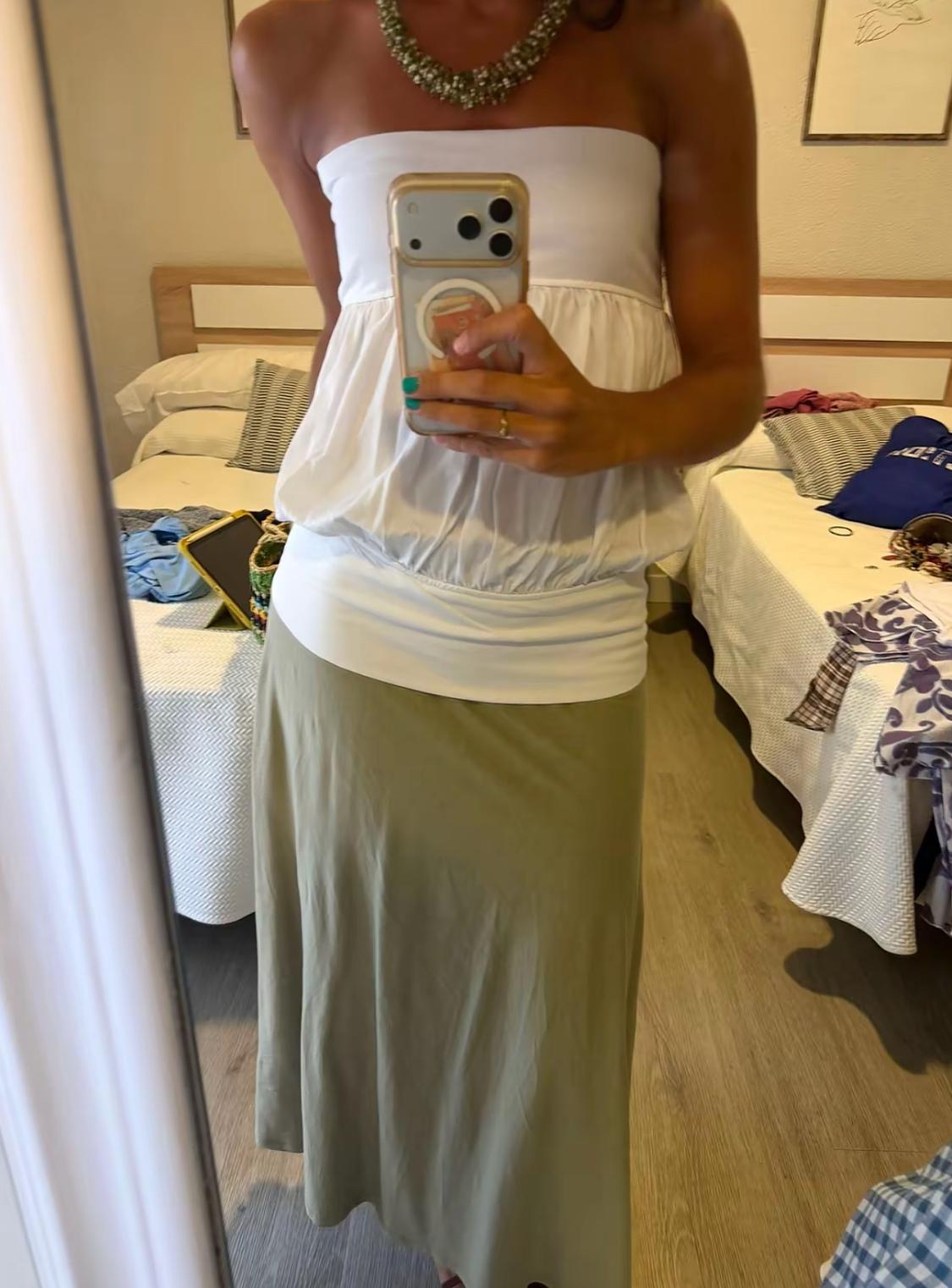 Mirror selfie of a woman in a bedroom. She is wearing a white strapless top, a long olive-green skirt and a chunky necklace.